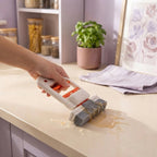 A person using a mini mop to clean a spill on a cream kitchen counter with dusty rose and lavender decor in the background.