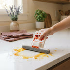 A hand using a compact mini mop to wipe a juice spill from a kitchen counter in a home setting.